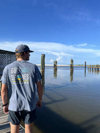 Person wearing a 'Gulf Shores, Alabama' t-shirt standing on a dock looking at the water.